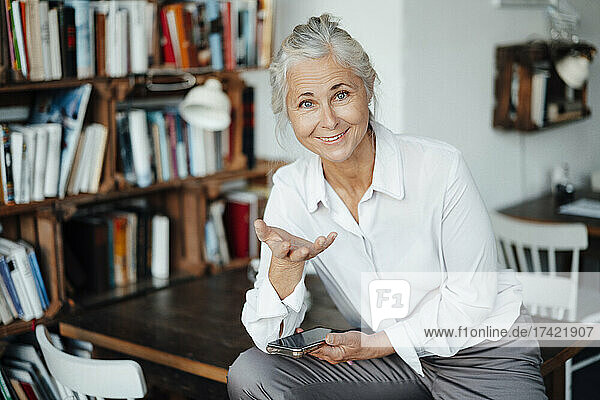 Female cafe owner with mobile phone gesturing while sitting on table