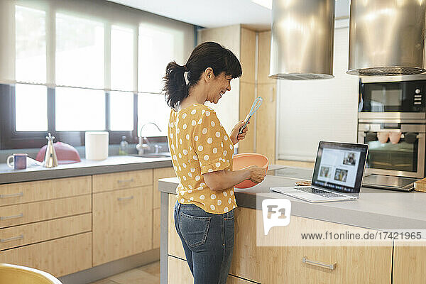 Glückliche Frau bereitet Essen zu  während sie in der heimischen Küche am Laptop lernt