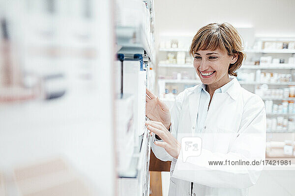 Smiling female pharmacist wearing lab coat searching medicine at pharmacy store