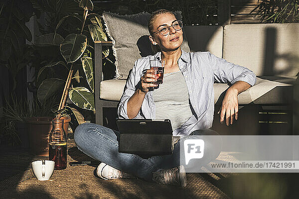 Thoughtful female freelancer holding drink with digital tablet on terrace