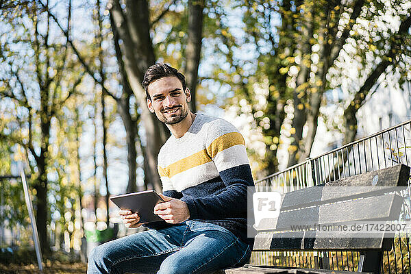 Smiling young man holding digital tablet while sitting on bench at park during sunny day