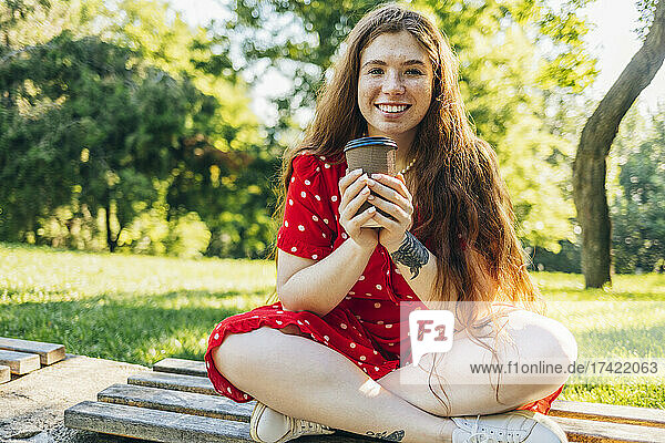 Young redhead woman with coffee cup sitting at park during summer
