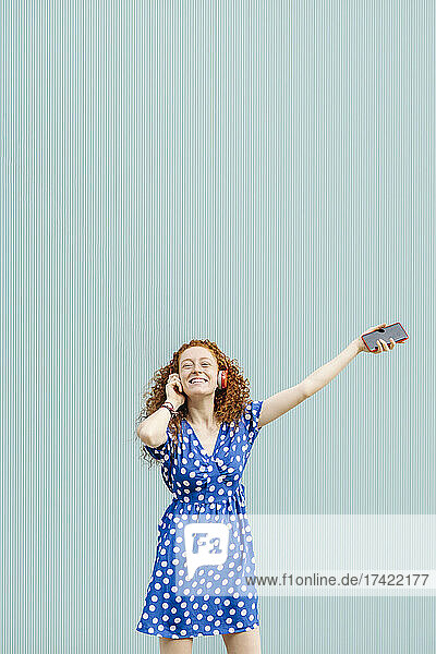 Smiling young redhead woman enjoying music in front of blue wall