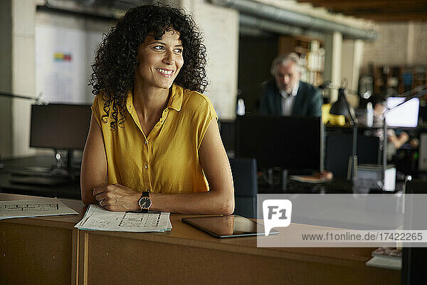 Smiling curly haired businesswoman with documents and digital tablet leaning on table at office