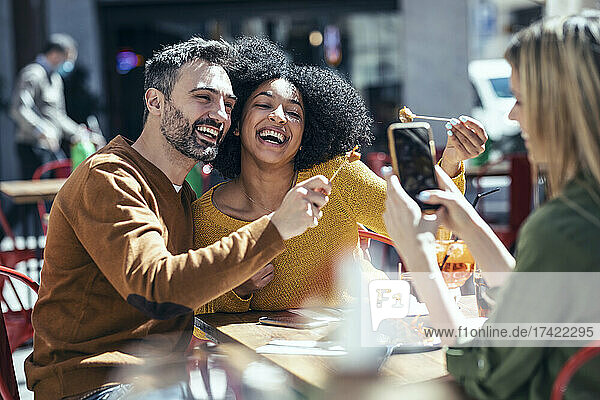 Woman photographing cheerful male and female friends showing food at restaurant