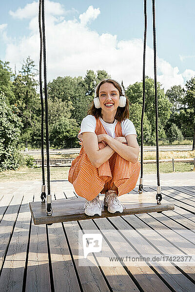 Smiling woman with headphones crouching on swing at park