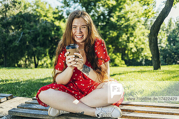 Cheerful woman holding disposable coffee cup while sitting with cross-legged on bench