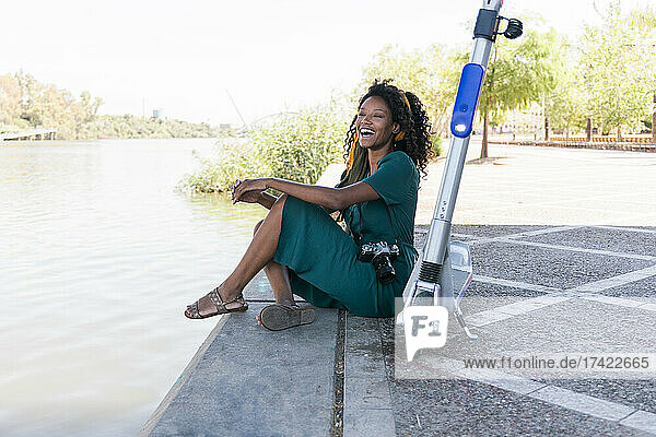 Cheerful Afro woman sitting with electric push scooter at riverbank