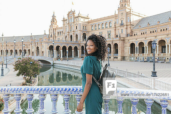 Lächelnde junge Frau mit lockigem Haar steht am Geländer an der Plaza De Espana  Sevilla  Spanien