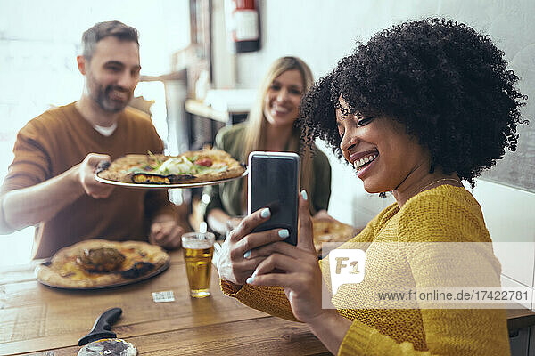 Happy Afro woman taking selfie with male and female friends having pizza in restaurant