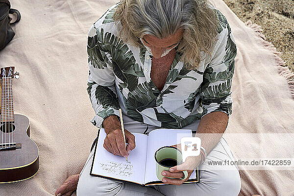 Mann mit Kaffeetasse schreibt in Buch  während er am Strand sitzt