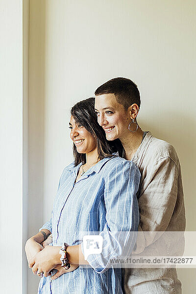 Lesbian couple looking away while embracing near wall at home