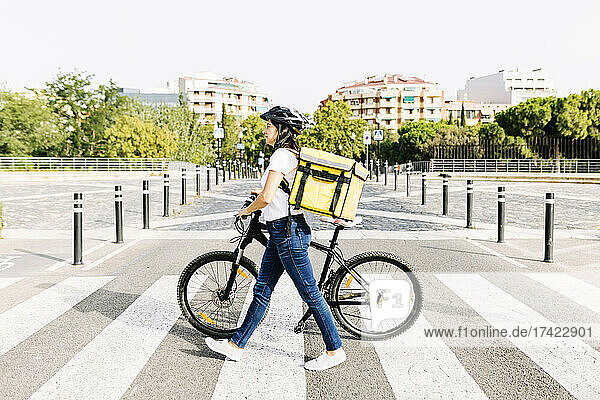 Delivery woman with backpack wheeling bicycle on road during sunny day