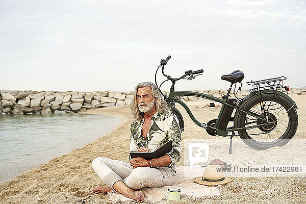 Man writing in book while sitting by bicycle at beach