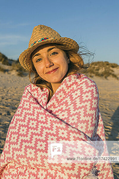 Smiling woman wearing sun hat wrapped in blanket at beach