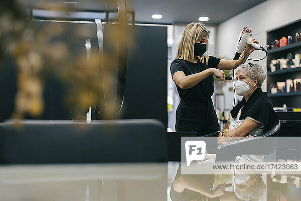 Female hairdresser with face mask blow drying customer's hair in salon