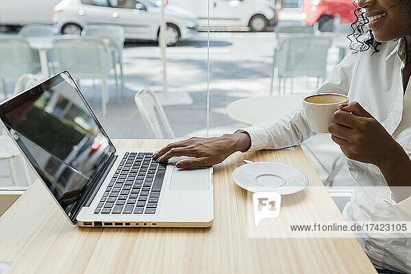 Female freelancer holding coffee cup while using laptop at cafe