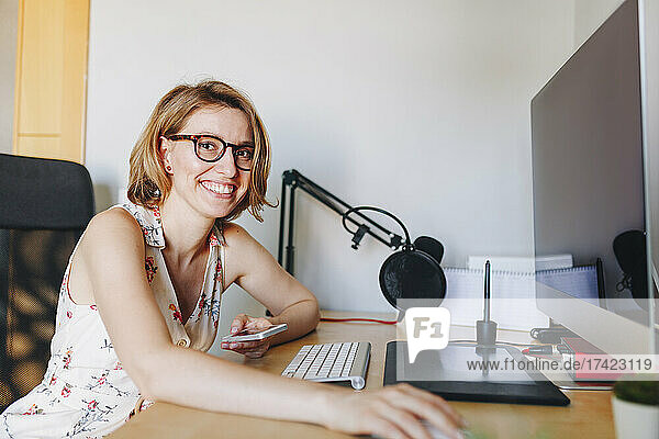 Smiling businesswoman holding mobile phone while sitting at computer desk in home office
