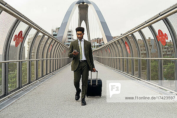Junger Geschäftsmann nutzt Mobiltelefon  während er mit Rollgepäck auf der Brücke läuft