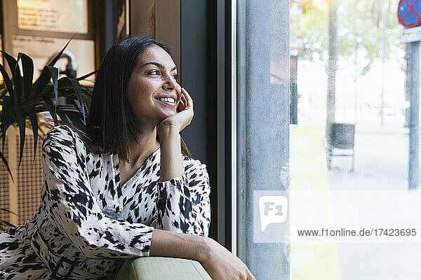 Smiling young woman day dreaming while looking through window in coffee shop