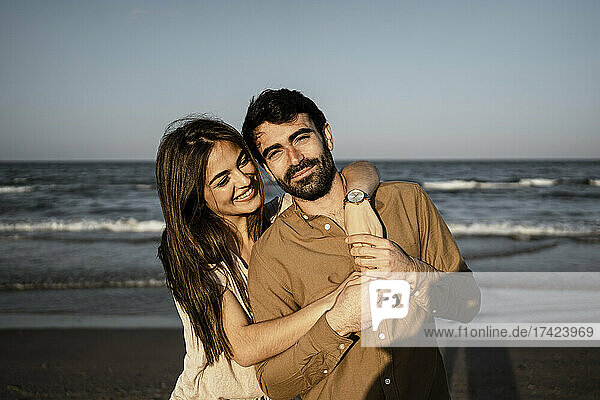 Lächelnde Frau umarmt Mann von hinten am Strand
