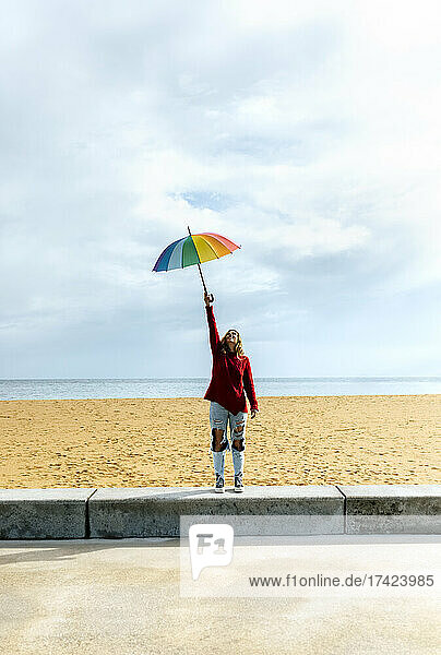 Woman holding umbrella with hand raised while standing in front of cloudy sky at beach