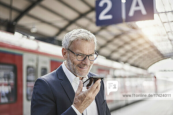 Smiling businessman sending voicemail through mobile phone at railroad platform