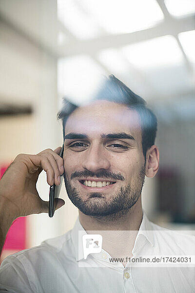 Lächelnder junger Geschäftsmann  der durch Glas auf einem Smartphone spricht