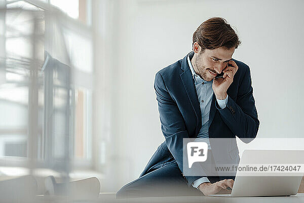 Young businessman using laptop while talking on mobile phone in office