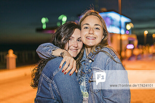 Smiling female friends in denim jackets embracing at night