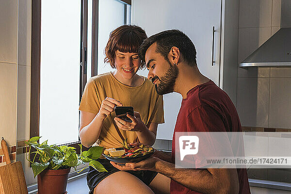 Lächelnde Frau fotografiert Essen  das ihr Freund zu Hause in der Küche hält