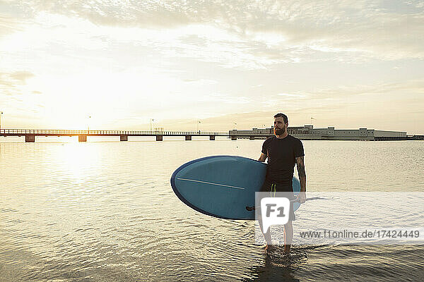 Mann schaut weg und trägt Paddleboard im Meer bei Sonnenuntergang