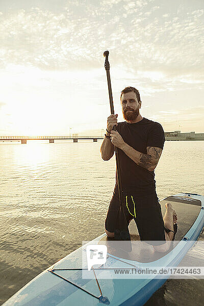Portrait of man holding oar while kneeling on paddleboard in sea