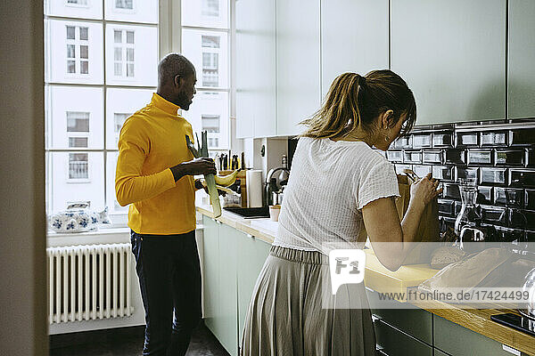 Mittlere erwachsene Frau  die Lebensmittel aus dem Papier nimmt  während ihr Freund mit Gemüse in der Küche zu Hause steht