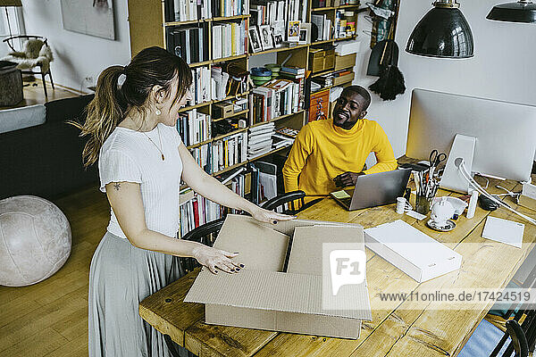 High angle view of male freelancer looking at happy girlfriend opening box standing at table