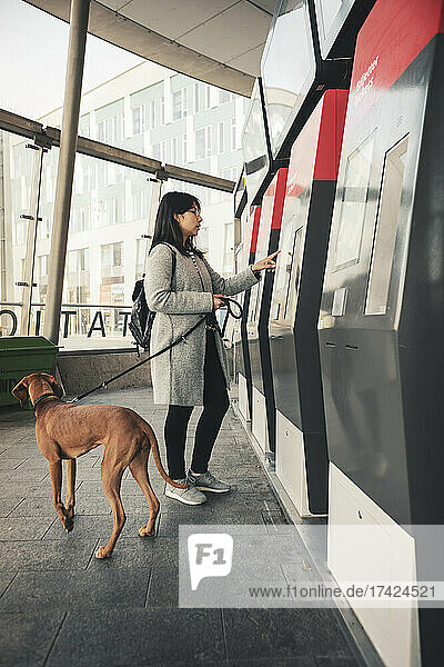 Junge Frau benutzt einen Fahrkartenautomaten  während sie mit ihrem Hund am Bahnhof pendelt