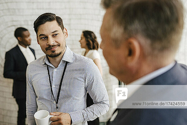 Lächelnder männlicher Unternehmer mit Blick auf einen reifen Geschäftsmann  der während eines Seminars im Kongresszentrum diskutiert