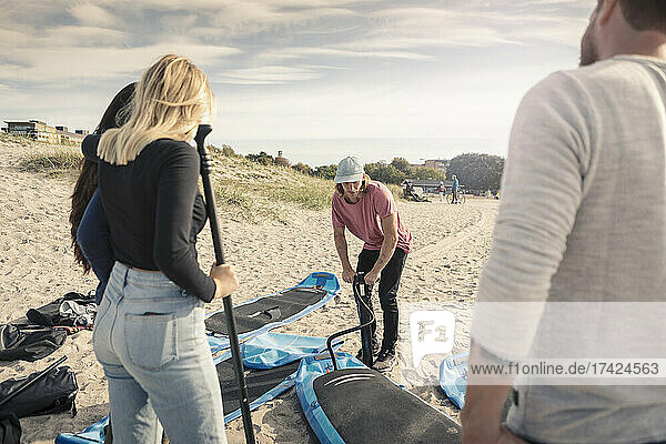 Mann pumpt Stand Up Paddleboard  während Freunde am Strand stehen