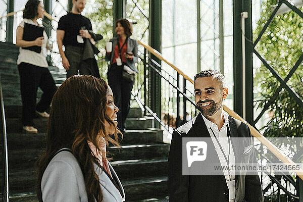 Lächelnde männliche Fachkraft im Gespräch mit einer weiblichen Kollegin während einer Networking-Veranstaltung  während sie an der Treppe steht