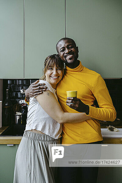 Porträt eines lächelnden Paares  das in der Küche zu Hause steht und den Arm um sich legt