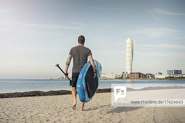 Rear view of man walking with stand up paddleboard at beach