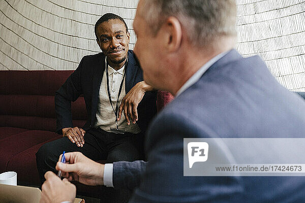 Smiling businessman looking at male colleague discussing while sitting on sofa during networking event
