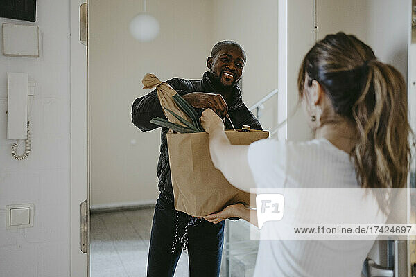 Ein lächelnder Mann gibt einer an der Tür stehenden Frau eine Papiertüte mit Lebensmitteln