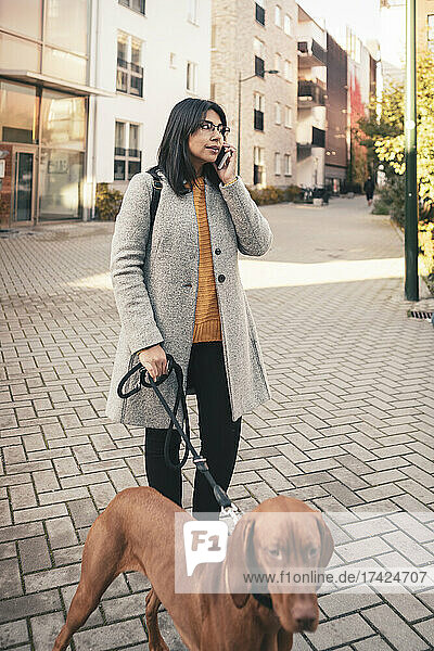 Junge Frau spricht in ihr Smartphone  während sie mit ihrem Hund auf dem Gehweg spazieren geht