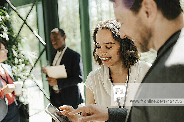 Lächelnde junge Geschäftsfrau  die sich mit einem männlichen Kollegen über ein Smartphone bei einer Networking-Veranstaltung unterhält