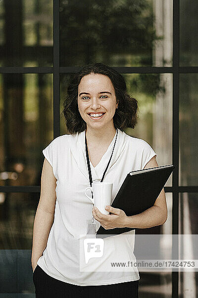Porträt einer glücklichen jungen Geschäftsfrau  die mit der Hand in der Tasche vor einer Glaswand im Kongresszentrum steht