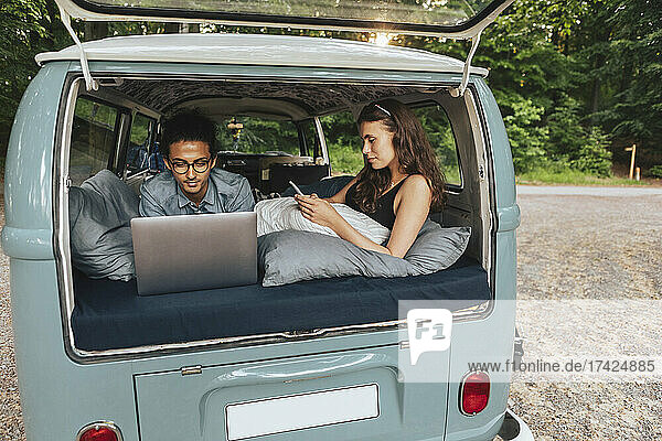 Mann mit Laptop  während Freundin mit Smartphone im Urlaub im Van liegt