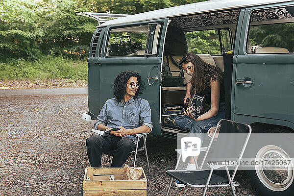 Junge Frau spielt Gitarre  während ein männlicher Freund mit einem Buch neben einem Van sitzt