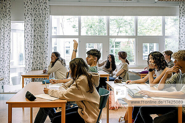 Junge hebt die Hand  während er mit anderen Schülern im Klassenzimmer sitzt