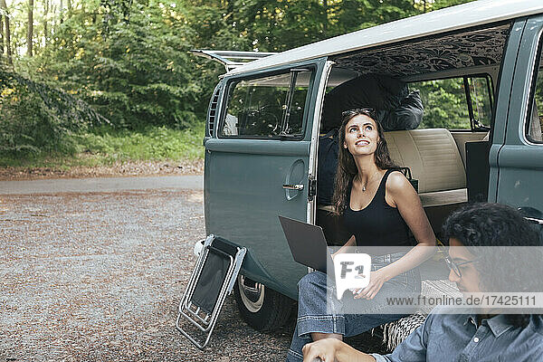 Frau mit Laptop und Blick nach oben  während sie neben einem männlichen Freund an einem Van sitzt